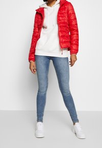 Rød pufferjakke med hætte og lynlås, båret over en hvid hættetrøje. Kombineret med lyseblå tætsiddende jeans og hvide sneakers.