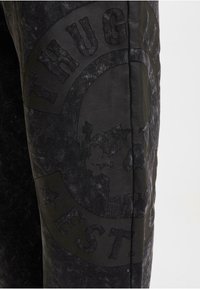 Svarta byxor med en texturerad finish och en upphöjd grafisk design, inklusive "THUG LIFE" och ett skalle, accentuerad med stjärnor.