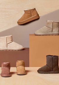 Quatre paires de bottines en daim dans des couleurs tan, beige, rose et marron, avec doublure en shearling, exposées sur un présentoir géométrique sur un fond en bois.