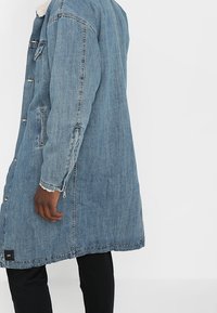 Abrigo largo de denim en azul claro con cuello de piel sintética, grandes bolsillos laterales, cierres con botones metálicos y diseño de corte recto.