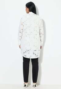 Camicia bianca a maniche lunghe con motivo a fiori ritagliato, polsini con bottoni, orlo asimmetrico, abbinata a pantaloni neri e tacchi bianchi.
