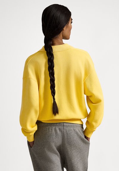 Kobieta z długimi splecionymi warkoczami, ubrana w żółty sweter i szare spodnie dresowe, stojąca z rękami w kieszeniach, zwrócona tyłem do kamery.