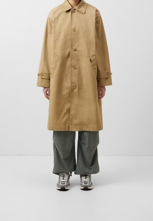 Personne portant un trench-coat beige longueur genou avec fermeture à boutons, pantalon gris à jambes larges et chaussures de sport grises, debout devant un fond uni.