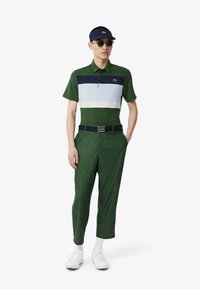 Grön pikétröja med vita och marinblå ränder, kombinerad med gröna mönstrade byxor och vita skor. Tillbehör inkluderar solglasögon och en skärm.