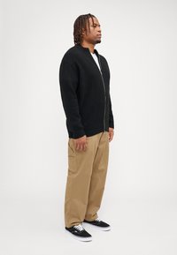Sort zip-up cardigan med en ribbet krave, iført over en hvid skjorte. Sættes sammen med khaki-bukser og sorte sneakers med hvide såler.