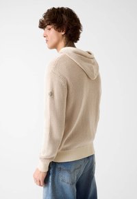 Maglione beige lavorato a maglia con cappuccio, caratterizzato da un motivo a rilievo, polsini e orlo a coste, e una discreta toppa con logo sulla manica sinistra.