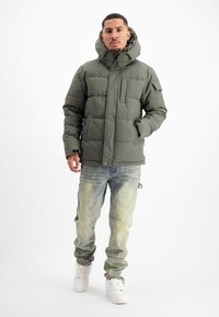 Gevoerd groen jack met een hoge kraag, rits- en knoopsluiting, grote capuchon en twee zijzakken, gecombineerd met vervaagde blauwe spijkerbroek en witte sneakers.