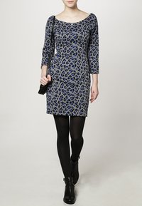 Robe à imprimé léopard gris avec des manches trois-quarts, des accents bleus et une silhouette ajustée ; portée avec des collants noirs et des bottines.