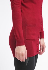 Roter Cardigan aus weichem Stoff, mit langen Ärmeln, einer Knopfleiste auf der Vorderseite und einer einzelnen Tasche auf der linken Seite. Strukturierte Bündchen.