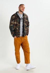Veste bomber camouflage avec des poignets côtelés, sweat-shirt gris avec logo et pantalon cargo orange. Des baskets blanches complètent le look.