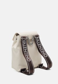 Krämfärgad ryggsäck med ett klafflock, silverkedjedetalj och breda mörkbruna remmar med tryckt "VALENTINO" i vitt.