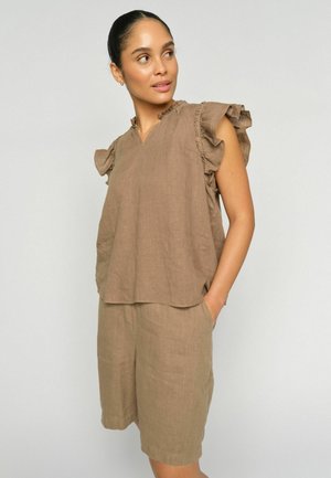 Žena odjevena u lepršavu bež majicu s volančićastim rukavima i odgovarajuće kratke hlače do koljena, stoji s jednom rukom u džepu.