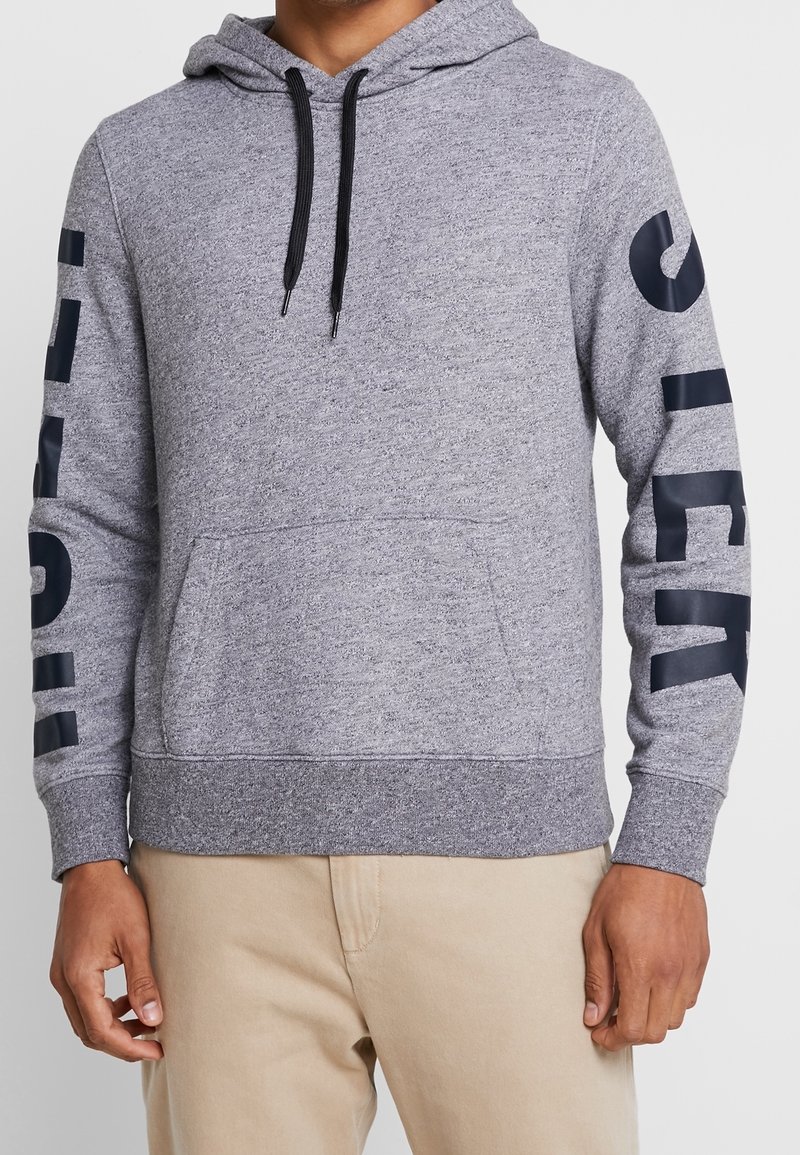 Grå hoodie med kontrasterande mörkblå text längs vänster ärm, tillverkad av blandmaterial av bomull, känguruficka och huva med snörning.