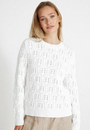 Witte gebreide trui met een golfpatroon, ribbelboord aan de ronde hals en zoom, en opengewerkte details voor textuur, gecombineerd met beige broeken.