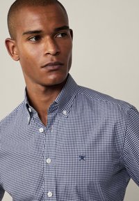 Blue and white checkered button-down shirt with a collar, featuring a small embroidered logo on the left chest and white buttons.