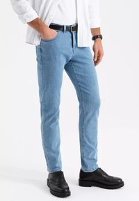 Muž ve světle modrých džínech, bílém tričku s ohrnutými rukávy, černým opaskem, černými společenskými botami a černými náramkovými hodinkami, stojící s jednou rukou v kapse.