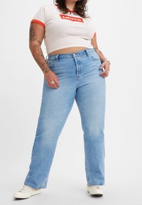 Jeans de tiro alto de color azul claro con un corte recto, combinados con una camisa corta blanca que presenta un logo rojo de Levi's y un ribete rojo en contraste.