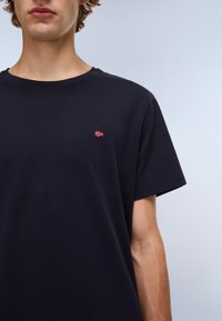 Marineblauwe katoenen T-shirt met een ronde hals en korte mouwen. Heeft een klein geborduurd rood-wit vlaggenembleem op de borst.