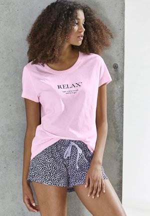 Woman with curly hair wearing a pink "RELAX" T-shirt and purple leopard-print shorts, standing against a concrete wall.