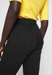 Jeans de mezclilla negra con un diseño ajustado, que cuentan con dos bolsillos traseros y acentos de costura visibles. Llevados con una blusa amarilla de canalé.