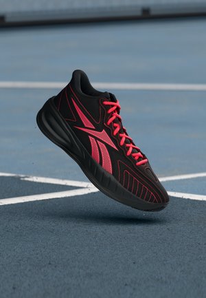 Scarpa sportiva nera e rossa sospesa sopra un campo esterno blu con linee bianche, che mostra il design dettagliato del lato e della suola.