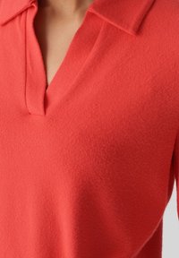 Haut corail éclatant en tissu doux et texturé, avec un col classique et un décolleté en V, présenté sous un angle pour mettre en valeur les détails du design.