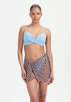 Kobieta ubrana w jasnoniebieski bikini zawiązywany na kopertę i przezroczysty brązowo-niebieski pasiasty sarong na tle jednolitej białej ściany.