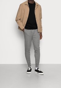 Beige kevyt takki mustan crewneck-paidan, harmaiden kapenevien jogger-housujen ja mustien valkoisilla pohjilla olevien tennareiden kanssa, taustalla yksinkertainen tausta.