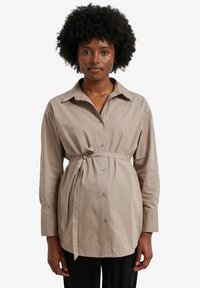 Beige button-up shirt with long sleeves, cinched waist tie, and collar. Smooth fabric with subtle texture and a relaxed fit.