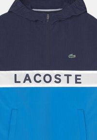Rompevientos ligero en navy y azul con cremallera de cuarto. Diseño a rayas. Presenta una banda blanca con el texto y logo de "LACOSTE".