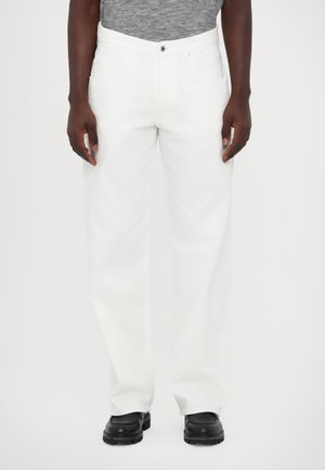 Personne portant un pantalon blanc droit, une chemise grise et des chaussures noires, debout devant un fond blanc uni.