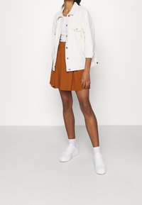 Veste en denim blanc portée sur un haut blanc, associée à une jupe évasée couleur rouille. La tenue comprend des baskets blanches épaisses et des chaussettes courtes.