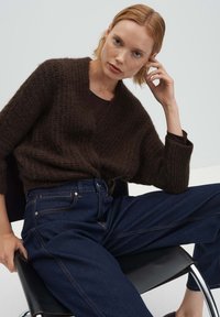 Donna con capelli biondi corti che indossa un maglione marrone scuro a maglia e jeans blu, seduta su una sedia nera contro uno sfondo semplice.