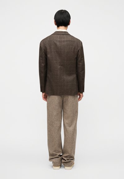 Καφέ, υφασμάτινο σακάκι με διακριτικό καρό σχέδιο, συνδυασμένο με φαρδιά μπεζ παντελόνια. Η εμφάνιση έχει έναν χαλαρό και μοντέρνο σχεδιασμό.