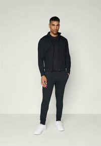 Jacket desportivo preto com fecho zip sobre uma camisa preta justa e calças slim-fit combinando, acompanhados de sapatilhas brancas. Tecido liso e respirável.
