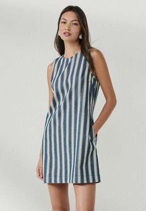 Junge Frau trägt ein ärmelloses blau-weiß gestreiftes Kleid mit vertikalen Streifen und seitlichen Taschen, steht vor einem schlichten hellen Hintergrund.