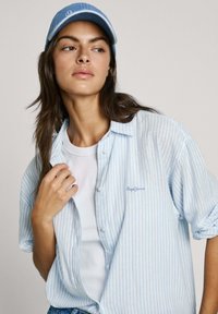 Hellblau gestreifte, locker sitzende Bluse mit kurzen Ärmeln. Darunter befindet sich ein weißes Tanktop und eine blaue Kappe.
