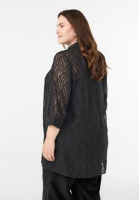 Žena s dlhými hnedými vlasmi, oblečená v čiernej priehľadnej blúzke s dlhými rukávmi a vlnitými vzormi, a čiernych nohaviciach, stojaca bokom na bielom pozadí.