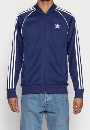Homme portant une veste de survêtement Adidas bleu marine avec des bandes blanches sur les manches et le logo sur la poitrine, assortie à un jean bleu clair.