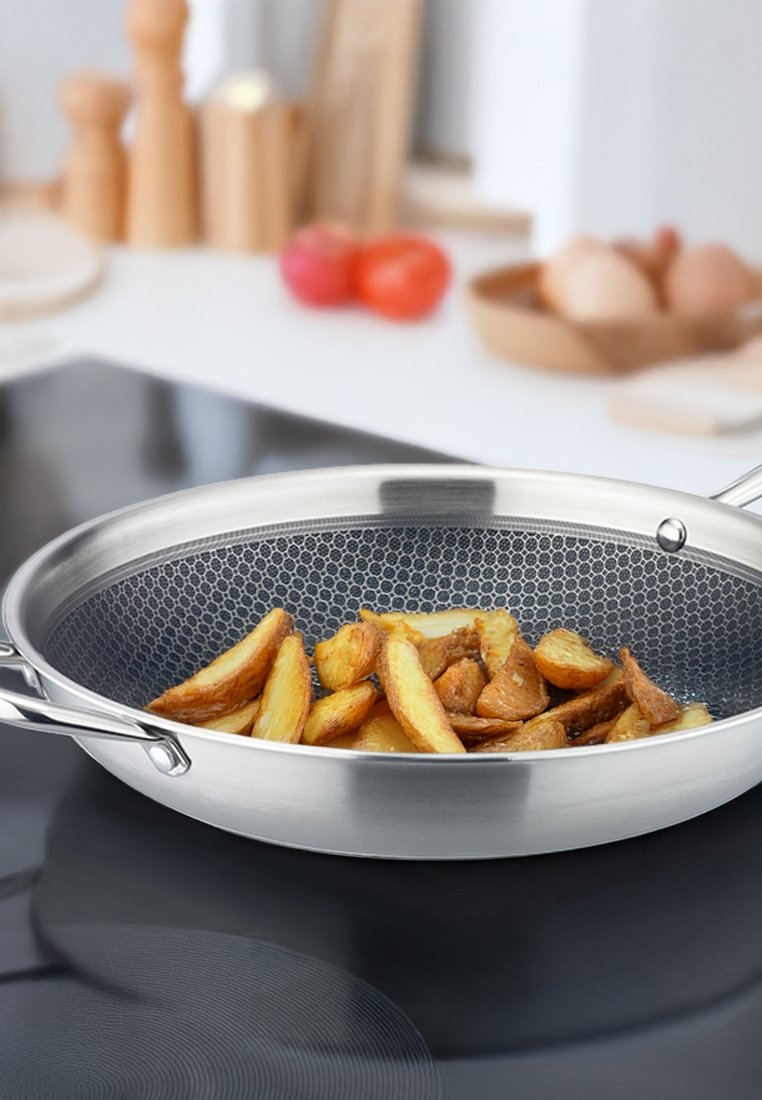 Edelstahlbratpfanne mit einem honigwabenmuster im Inneren, gefüllt mit goldbraun gebratenen Kartoffeln, vor einem modernen Küchenhintergrund.