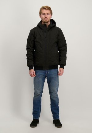 Zwarte geïsoleerde jas met een capuchon, voorrits en elastische manchetten, gecombineerd met blauwe spijkerbroek en zwarte laarzen. Gladde textuur, casual ontwerp.