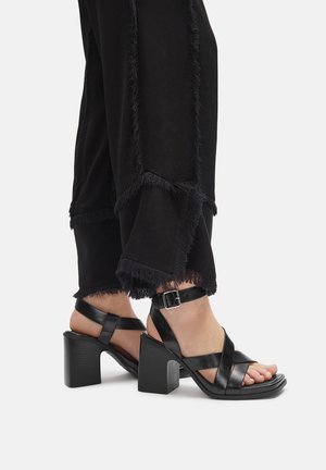 Fekete, csatos bokapántos, blokkjárgású szandálban lévő lábfejek, fekete, rojtos szegélyű, rövid nadrággal párosítva.