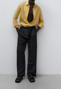 Camisa de lino amarilla, abierta en el cuello. Pantalones negros con pliegues y textura suave. Sandalias marrones con tiras ajustables.