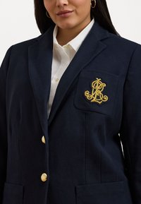 Marineblazer med strukturert stoff, med skrå lapels, gullknapper og brodert logo på brystlommen. Hvit skjorte under.