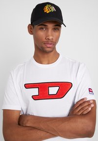 White cotton t-shirt with red textured "D" logo, short sleeves, and black cap featuring an embroidered colorful Native American face design.