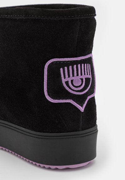 Botín negro de ante que presenta un diseño de ojo bordado en púrpura en el costado, con una suela de goma texturizada y un acabado suave.