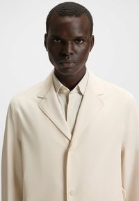 Blazer beige in tessuto texturizzato, con colletto a revers e bottoni frontali, indossato su una camicia crema chiara con colletto abbottonato.