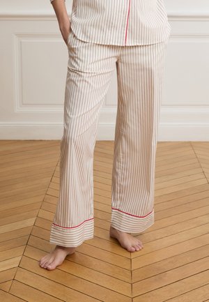 Persona descalza de pie sobre un suelo de madera, vistiendo pantalones sueltos de color crema con finas rayas verticales y ribete rojo cerca de los puños.