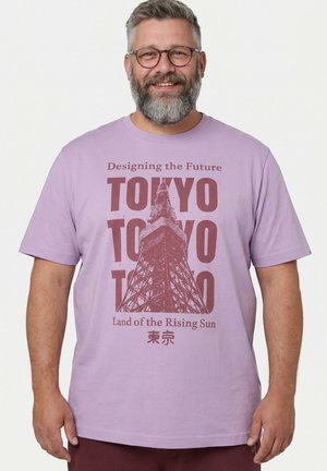 Lächelnder Mann mit Brille und Bart trägt ein hellviolettes T-Shirt mit einer Grafik des Tokyo Tower und einem Text über Tokyo und die aufgehende Sonne.