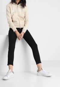 Persona seduta su un blocco bianco indossando un hoodie beige GANT, pantaloni neri aderenti e sneakers bianche con dettagli verdi.